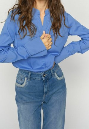 Vrouw met lang bruin haar die een blauw shirt met lange mouwen en een blauwe spijkerbroek draagt, met één hand op haar borst en de andere op haar heup.