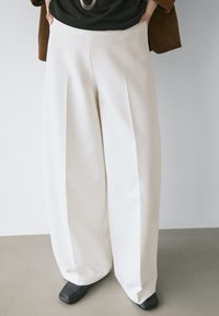 Personne portant un pantalon blanc à jambes larges, des chaussures noires à bouts carrés, un haut noir et une veste marron, debout sur un sol clair contre un fond uni.