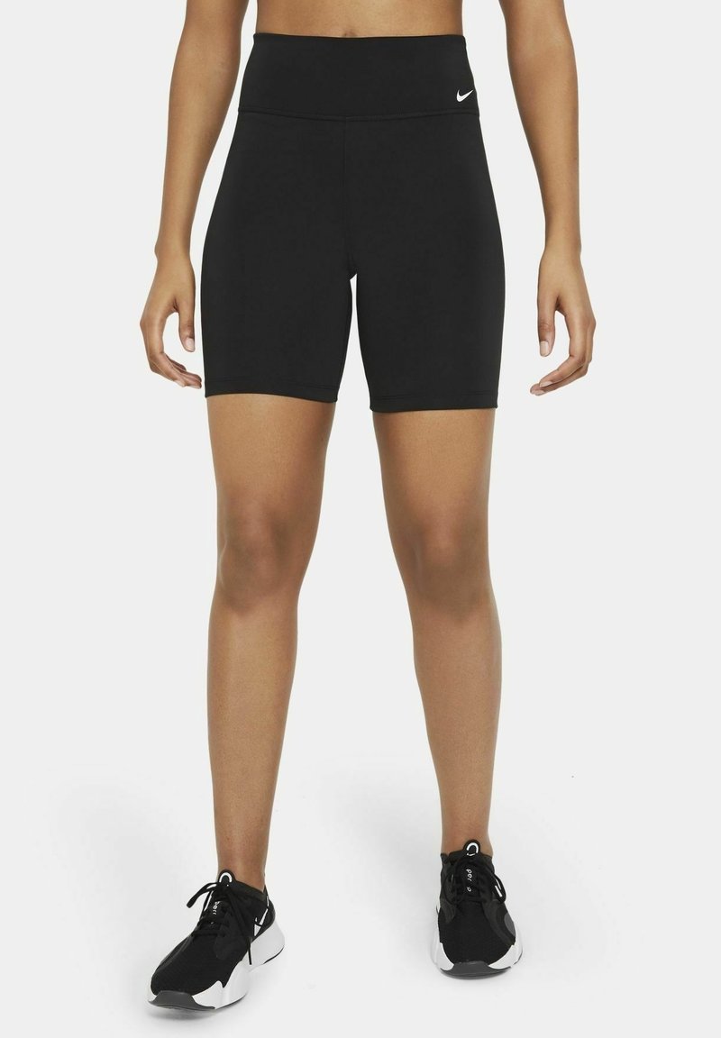 Personne portant un short de sport noir taille haute et des baskets noires et blanches, debout sur un fond uni.