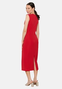 Vestido midi rojo sin mangas con un diseño elegante, abertura lateral y tela suave. La modelo lleva sandalias plateadas con tiras.