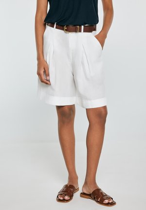 Kobieta w białych plisowanych szortach, brązowym skórzanym pasku, czarnym topie i brązowych płaskich sandałach, stoi z rękami w kieszeniach.