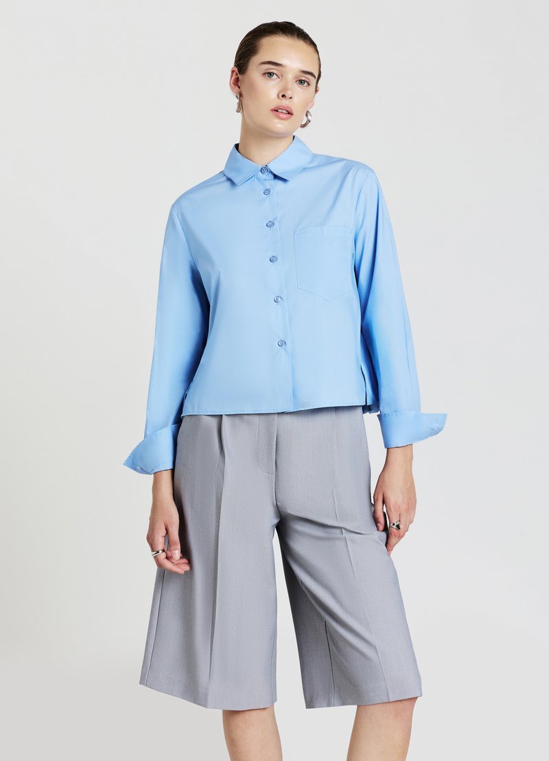 Hellblaues, langärmeliges Hemd mit Knopfleiste und Brusttasche, kombiniert mit grauen, knielangen, maßgeschneiderten Shorts. Glatter Stoff, lockere Passform.