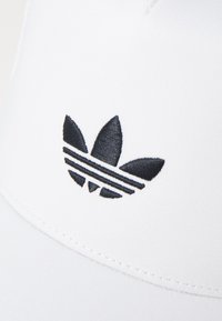 Valkoinen lippalakki, jossa on musta brodeerattu Adidas-logo, jossa on kolme lehteä ja kolme raitaa. Lähes sileä pinta ja kaareva lippa.