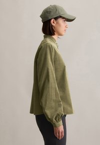 Chemise en velours côtelé vert olive à coupe décontractée, avec un col boutonné et des manches ballon, assortie d'une casquette. Des leggings noirs complètent le look.