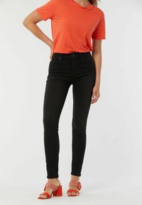 Jeans skinny noirs associés à un t-shirt orange vif et des sandales à talons blocs rouges avec une bride texturée.