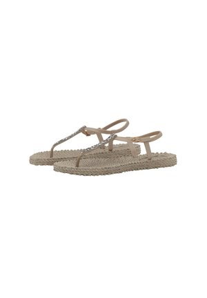 Sandalias planas beige con suelas trenzadas, parte superior de tira delgada en T adornada con piedras brillantes y cierre de hebilla ajustable en el tobillo.