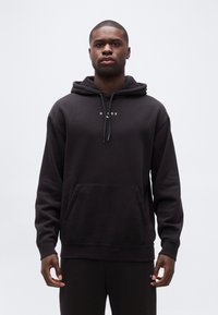Homme portant un sweat à capuche noir uni avec un petit logo blanc et un pantalon noir assorti, debout devant un fond clair neutre.