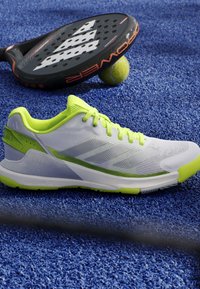 White and neon green sports shoe on blue tennis court surface with black and orange padel racket and yellow tennis ball.
