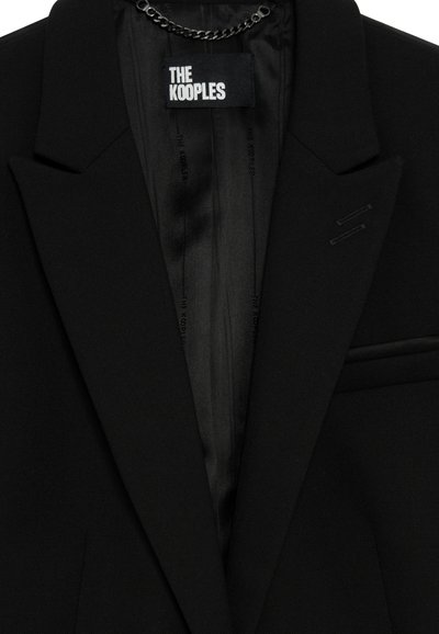 Blazer noir sur mesure avec revers crantés, poche passepoilée simple et étiquette intérieure visible portant la mention "THE KOOPLES."