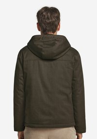 Mand iført en mørk olivengrøn hættetrøje, stående med ryggen mod beskueren foran en ensfarvet hvid baggrund.