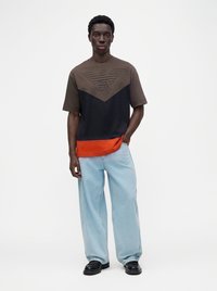 Jeune homme portant une chemise à manches courtes color block marron, noir et orange, et un jean bleu clair ample avec des chaussures noires.