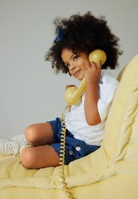 Gele vintage telefoonhoorn, kind met krullend haar en blauwe strik, draagt een wit shirt en spijkerbroekshorts, zittend op een gele bank.