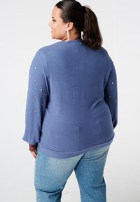 Vrouw met donker haar in een paardenstaart, gekleed in een blauwe trui met pareldetails op de mouwen en lichtblauwe spijkerbroek, die glimlacht en naar links kijkt.