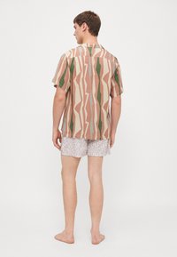 Camisa de manga corta con un patrón geométrico en beige, verde y rosa. Llevada sobre pantalones cortos gris claro con un diseño de logo sutil.