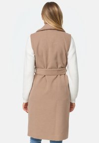Longue veste tan avec un large col, dotée d'une taille ceinturée et de manches longues blanches. Texture douce, design uni et coupe mi-longue.