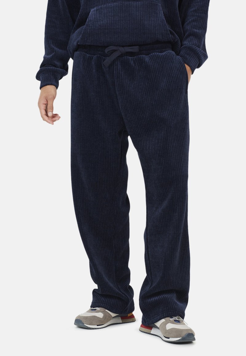 Mężczyzna noszący luźne granatowe spodnie kordowe i szare sneakersy z beżowymi i czerwonymi akcentami, dłonie swobodnie opadające wzdłuż ciała.