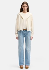 Felpa beige chiaro con zip e cappuccio, abbinata a una maglia aderente color crema e jeans dritti azzurri chiari. Tacchi marroni completano il look.