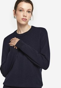 Granatowy sweter dzianinowy z długim rękawem i okrągłym dekoltem; miękka faktura; z prążkowanymi mankietami i brzegami. Model pozuje z rękami skrzyżowanymi.