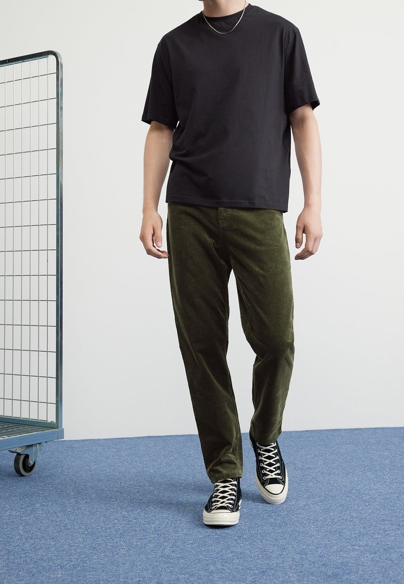 Zwarte korte mouwen t-shirt gecombineerd met groene corduroy broek en zwarte sneakers met witte accenten. Eenvoudige, relaxte pasvorm in casualkleding.