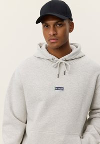 Φωτεινή γκρι φούτερ με ρυθμιζόμενο κορδόνι και μπροστινή τσέπη, με μπλε και πράσινο λογότυπο "LES DEUX". Φορεμένο με μαύρο καπέλο.