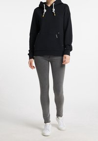 Sweat à capuche noir avec poche frontale, cordons de serrage avec accents jaunes, porté avec un jean gris et des baskets blanches. Texture lisse et coupe décontractée.