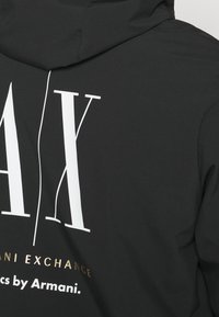 Svart huvtröja med en stor vit "AX"-logotyp. Har guldtexten "Armani Exchange" nedanför. Slät textur, avslappnad design och funktionell huva.