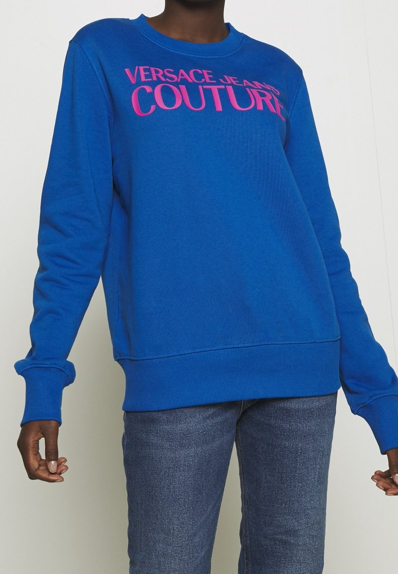 Personne portant un sweat-shirt bleu vif avec l'inscription « Versace Jeans Couture » en rose, associé à un jean bleu de lavage moyen.