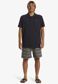 Marinblå polo med kort ärm, kombinerad med mönstrade svartvita shorts med zigzag-mönster och svarta sandaler.