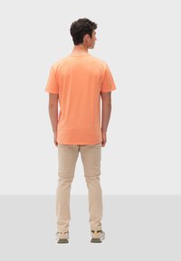 T-shirt in cotone arancione con maniche corte e scollatura rotonda, abbinata a pantaloni beige e sneaker grigie con dettagli verdi.