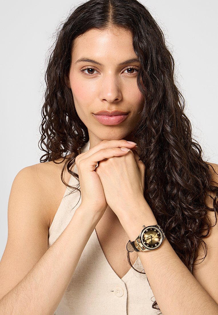 Reloj de pulsera en tono dorado con un dial redondo negro, bisel adornado y correa texturizada. Presenta un acabado pulido y números claros.