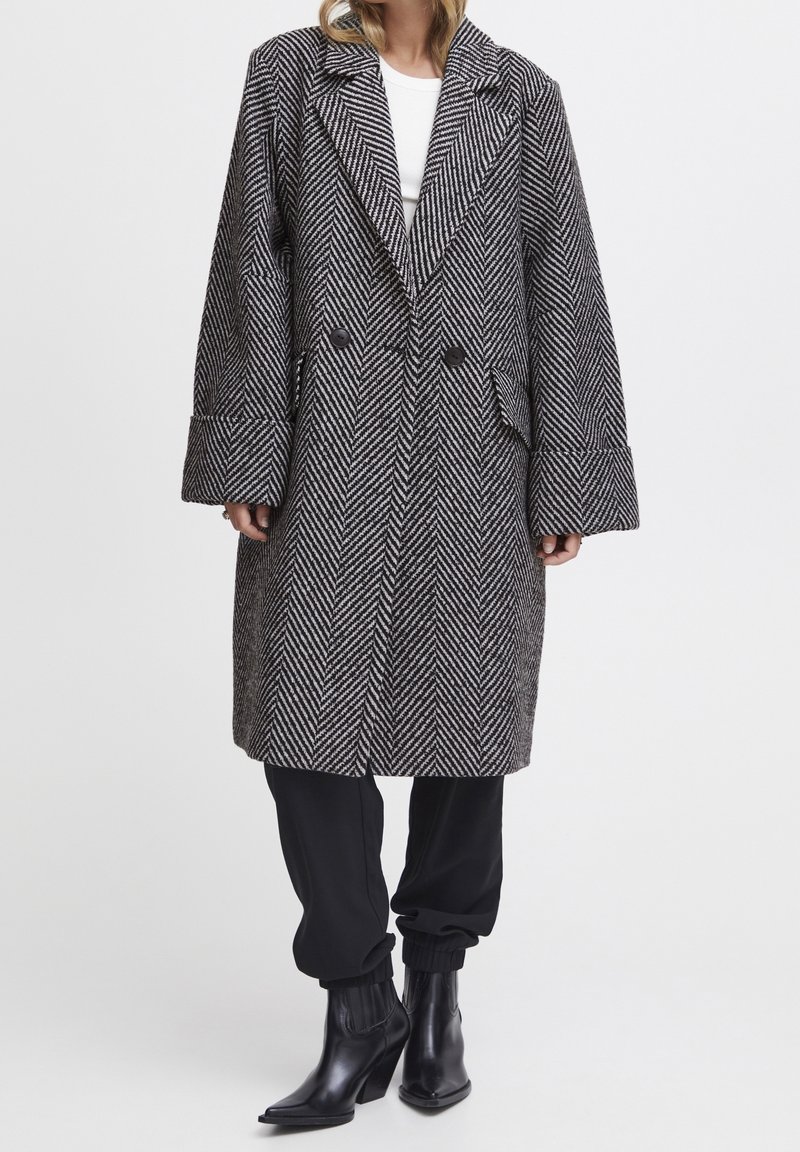 Femme portant un manteau croisé à motif chevrons noir et blanc, un pantalon noir et des bottines noires, sur un fond uni.