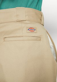 Pantaloni da lavoro beige con una texture liscia, dotati di una tasca posteriore e di una patch rettangolare con il logo Dickies. La cintura è cucita con una finitura pulita.