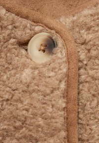 Tessuto fuzzy beige con una consistenza morbida, caratterizzato da un bottone rotondo di colore chiaro e da un bordo liscio a contrasto lungo il perimetro.