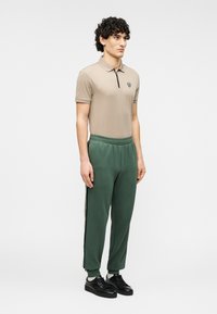 Μπεζ πόλο μπλουζάκι με μαύρο φερμουάρ και λογότυπο, σε συνδυασμό με πράσινη φόρμα με ρίγες στο πλάι. Ολοκληρώνεται με μαύρα αθλητικά παπούτσια.