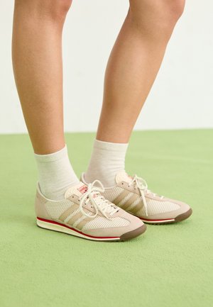 Beežid võrkketsid, millel on nubukaktsendid, kolm valget triipu, punane detail taldadel ja neutraalsed sokid, mis on kantud üle pahkluude.