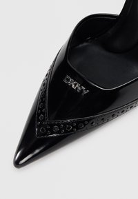 Čierna lakovaná kožená topánka na vysokom podpätku s špicatou špičkou, s perforovanými detailmi a strieborným logom "DKNY" na boku.