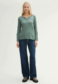 Groene langekuyper met een V-hals en knoopdetails, gecombineerd met wijdvallende donkerdenim jeans en grijze schoenen. Zachte, gestructureerde stof.
