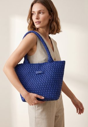 Femme portant une tenue beige sans manches tient un grand sac cabas bleu tissé étiqueté « DIANA&CO » sur son épaule.