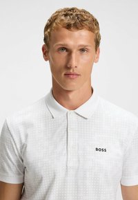 Vit polo tröja med ett subtilt geometriskt mönster, korta ärmar och klassisk krage. Varumärkeslogotypen "BOSS" i svart på bröstet.