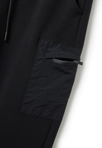 Schwarze Trainingshose mit einer kontrastierenden, strukturierten Stofftasche mit Reißverschluss. Glattes Material mit einer taillierten Passform. Keine sichtbaren Muster.