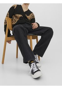 Cárdigan negro con patrón abstracto en tonos marrones, combinado con jeans negros holgados y zapatillas negras con suelas y cordones blancos, sentado en una silla de madera.