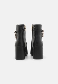 Bottines noires à talon épais, fabriquées en matériau synthétique lisse. Présente des accents de fermeture éclair dorés sur le côté extérieur.
