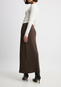 Longue jupe marron avec une texture lisse, assortie à un haut ajusté à manches longues blanc. Des bottes à talons noires complètent la tenue.