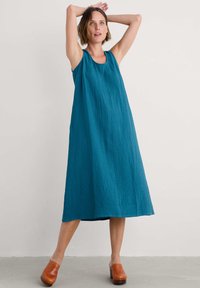 A sleeveless, loose-fitting dress in textured teal fabric, featuring a scoop neckline and falling to mid-calf, paired with brown wooden clogs.