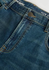 Slim-fit kavbojke v srednjem modrem pranju, z oranžnimi šivi, sprednjim gumbom za zapiranje in klasičnim pet-pocket dizajnom.