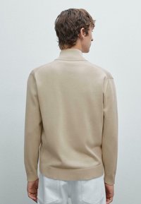 Beige, leichter Pullover mit geripptem Kragen und Bündchen, der eine glatte Textur und eine figurbetonte Passform aufweist, von hinten betrachtet.