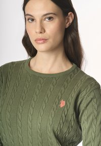 Una donna con i capelli lunghi indossa un maglione verde a tricot, con un piccolo emblema rosso sul petto, fissando direttamente la macchina fotografica.