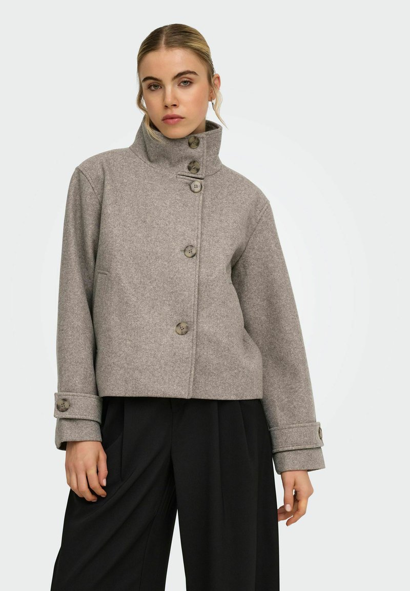 Femme portant une veste en laine grise boutonnée à col montant et un pantalon noir à jambes larges, debout devant un fond clair uni.