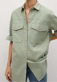 Chemise vert clair à boutons-pression en tissu doux, dotée de deux poches poitrine, manches retroussées et boutons en bois clair.