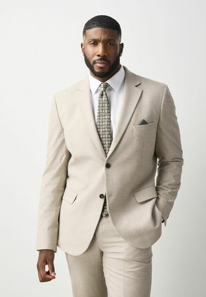 Man with beard wearing beige suit, white shirt, patterned tie, and pocket square stands with one hand in pocket against plain background.